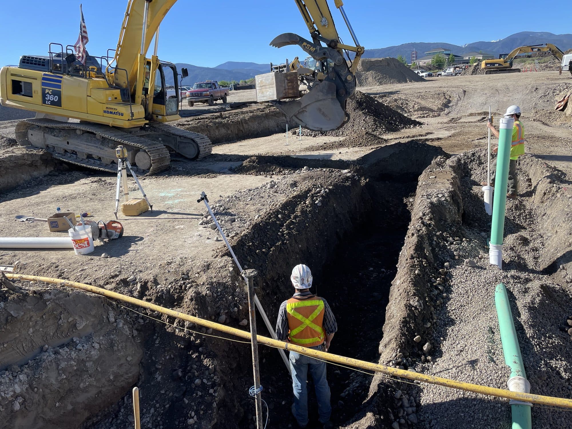 Excavator working over an open utility trench while a crew member checks the pipe run