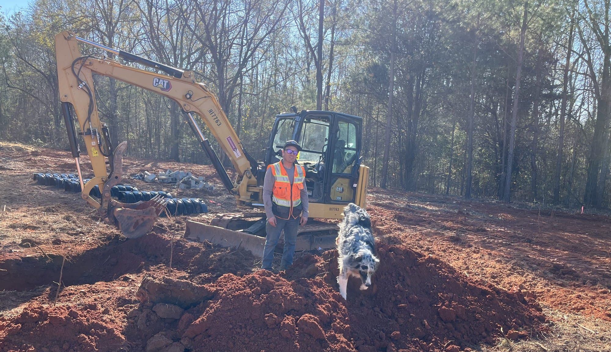 Joseph Caddel and his dog Bridger on a Carolina job site in front of a CAT mini excavator, with septic chambers staged nearby