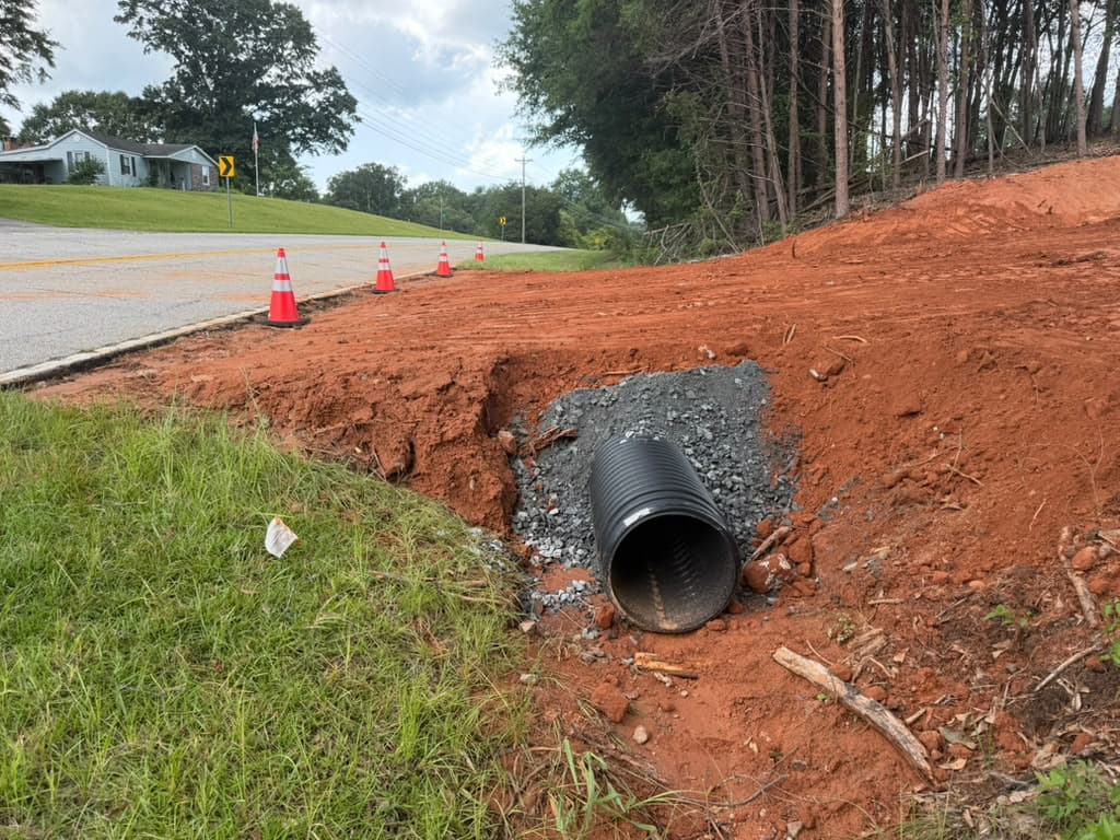 New culvert pipe bedded in gravel at a driveway entrance, with traffic cones set along the roadside