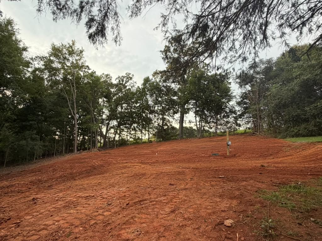 Sloping lot cleared and graded to final shape in red clay, with a utility post set near the treeline