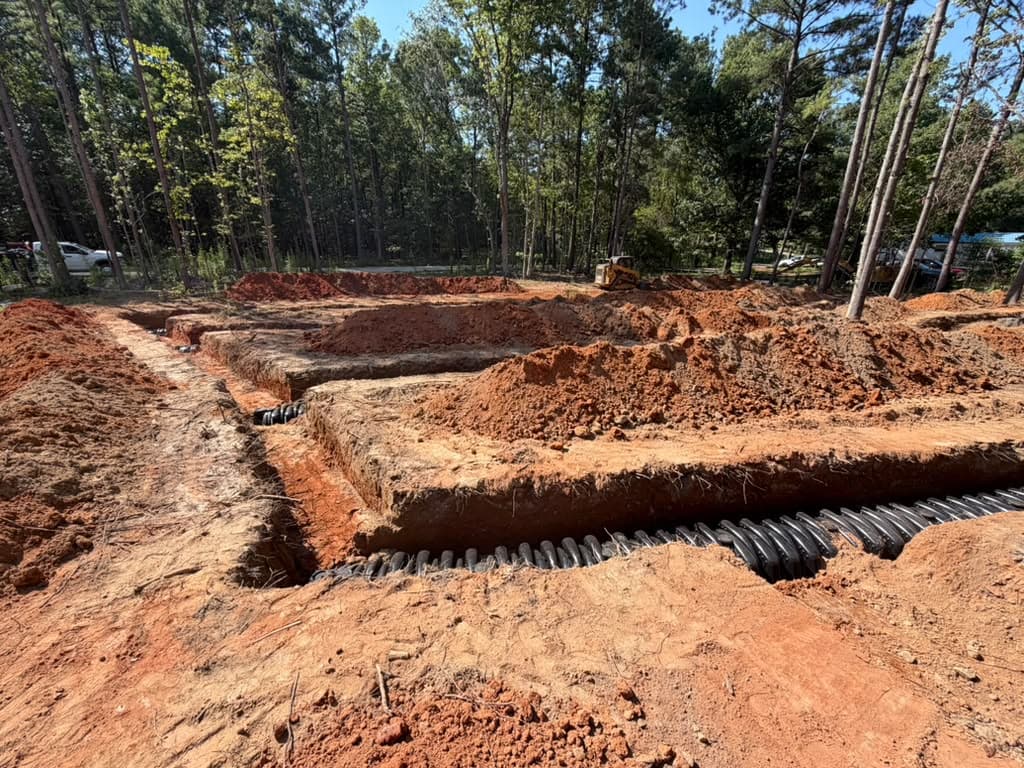 Corrugated drain field pipe laid in open septic trenches on a wooded lot, with a dozer working in the background