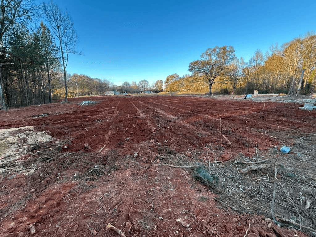 Cleared and grubbed lot in red Carolina clay, root debris raked to the treeline, ready for grading