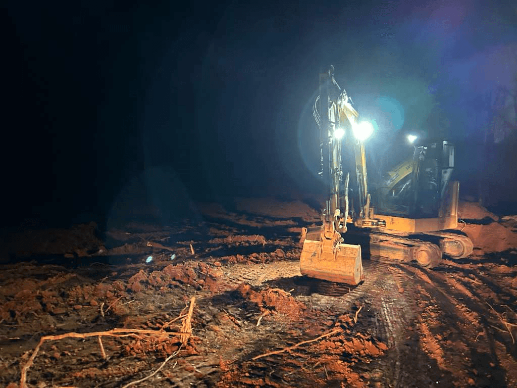 Excavator working under floodlights at night on freshly cleared ground strewn with roots and stumps